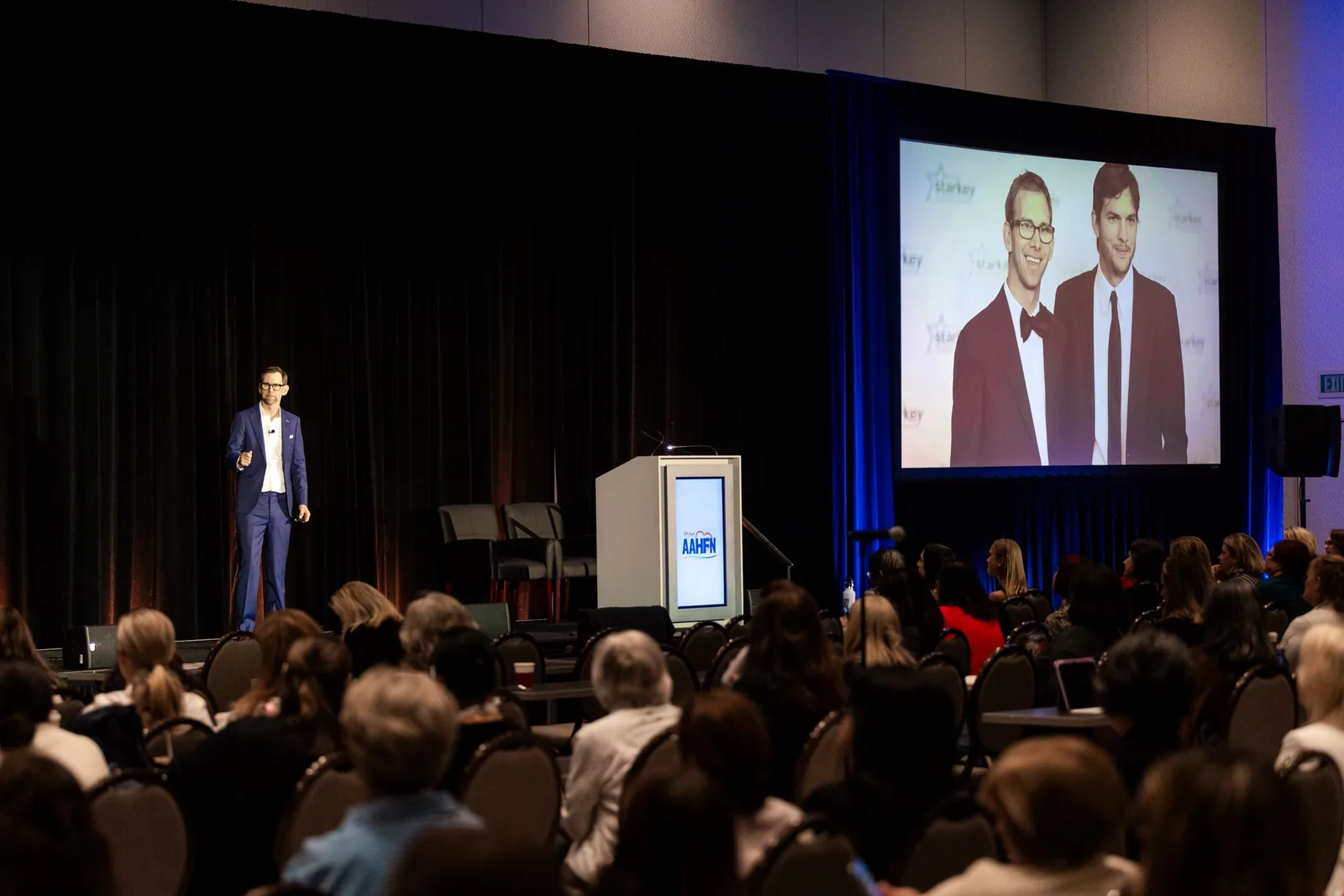 The room from the back, both screens carrying Michael Kutcher
