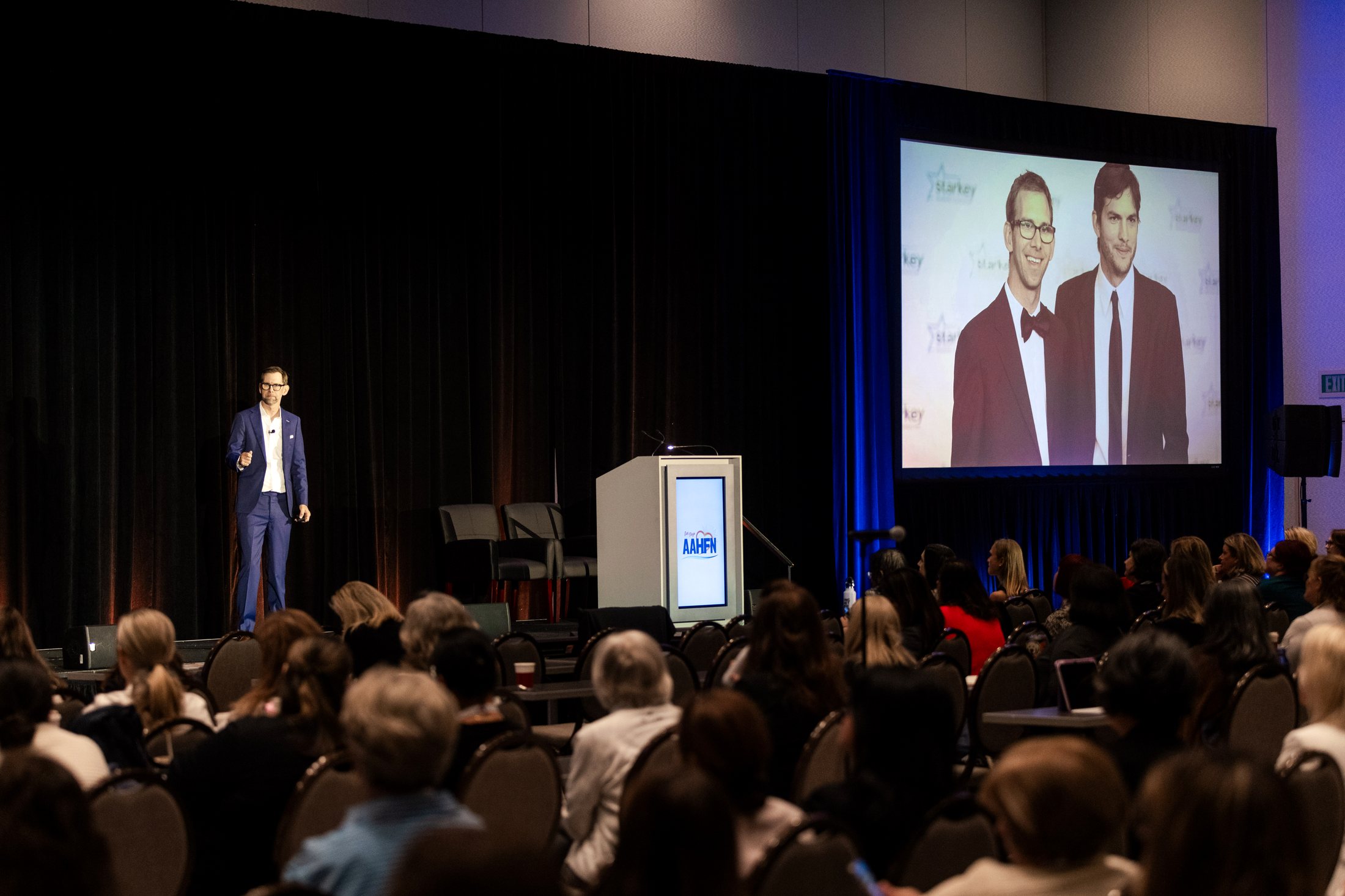 The room from the back, both screens carrying Michael Kutcher
