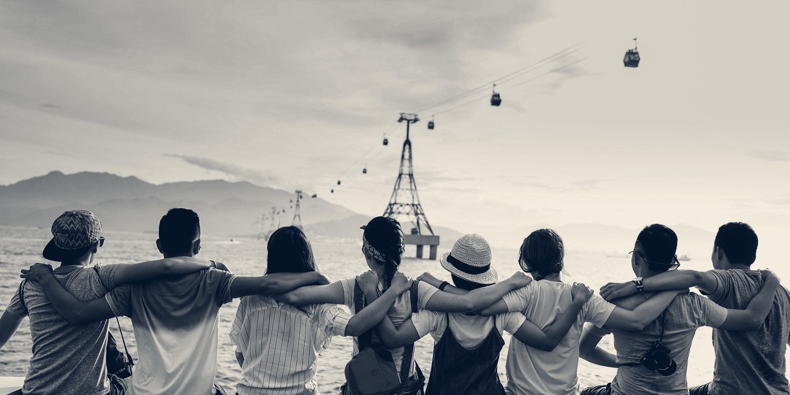 A row of people seen from behind, arms around each other's shoulders, looking out over water