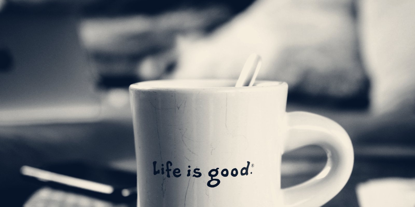 A white mug printed with the words Life is good, a blurred desk behind it