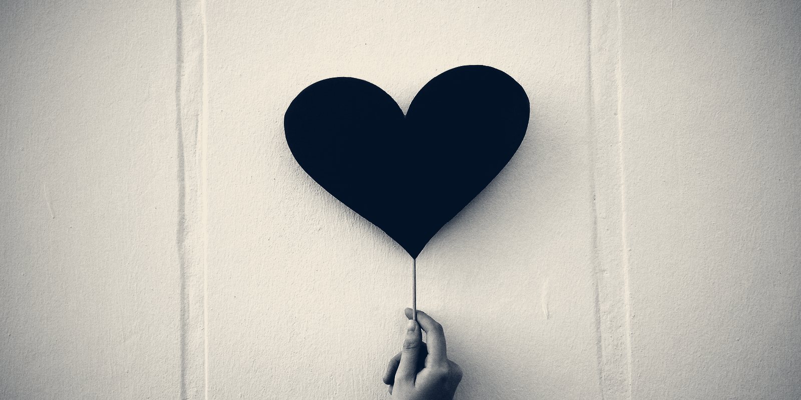 A hand holding up a dark paper heart on a stick against a pale wall