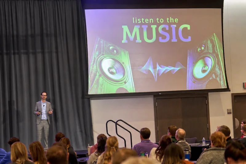 Michael Kutcher on stage beside a slide reading Listen to the Music, an audience seated in front
