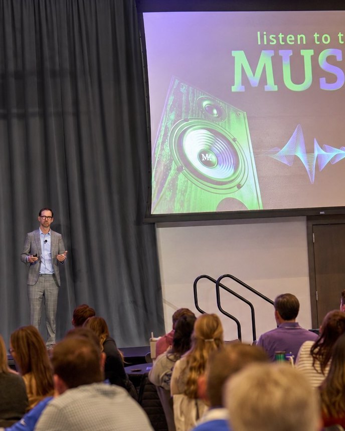 Michael Kutcher on stage beside a slide reading Listen to the Music