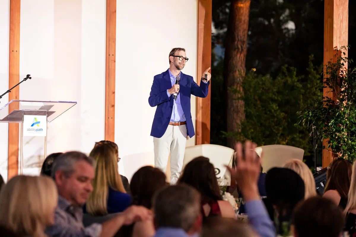 Michael Kutcher speaking into a handheld microphone at an outdoor evening event, the audience in the foreground