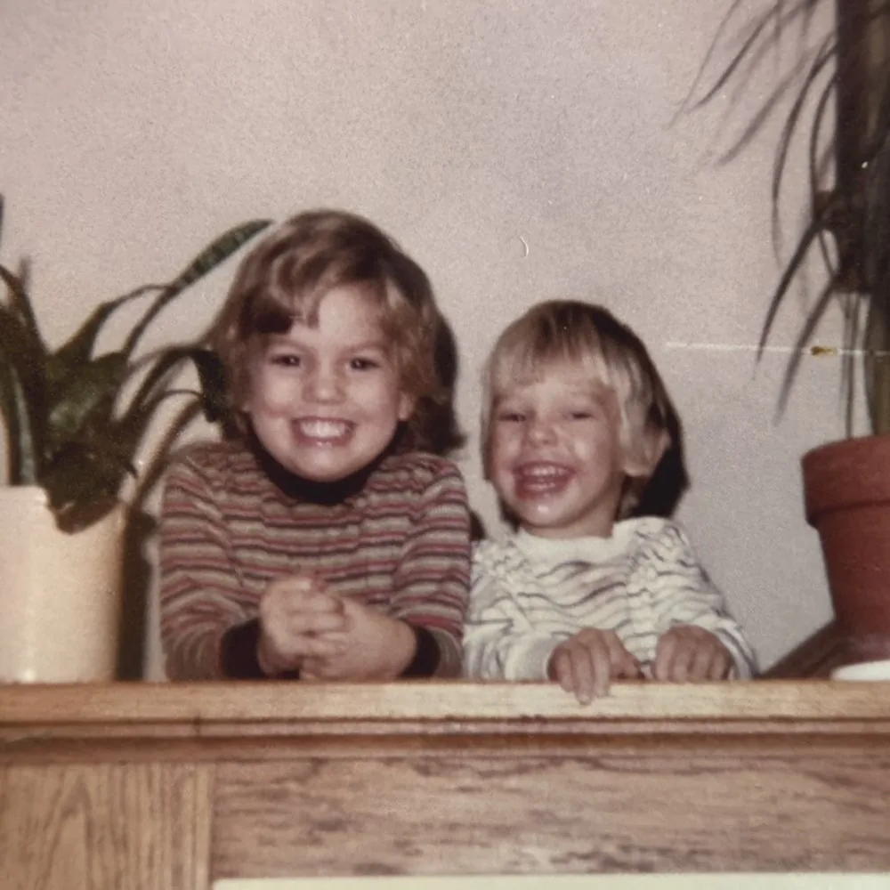 Two young children laughing behind a wooden mantel, with houseplants on either side