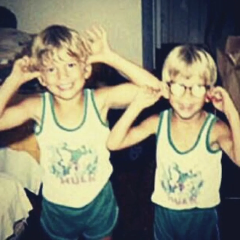 Two young boys in matching vests and green shorts pulling faces, one wearing glasses