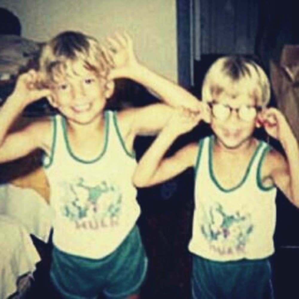 Two young boys in matching vests and green shorts pulling faces, one wearing glasses
