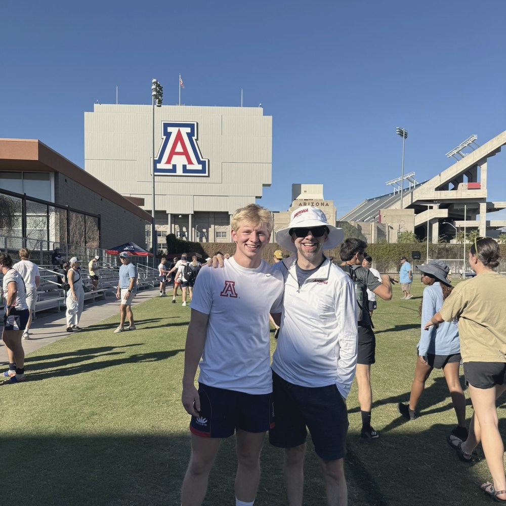 Two people standing arm in arm on a sunny field outside a stadium marked with a large letter A