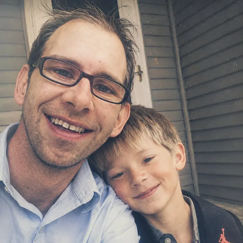 A man in glasses and a young boy leaning together and smiling outside a grey clapboard house