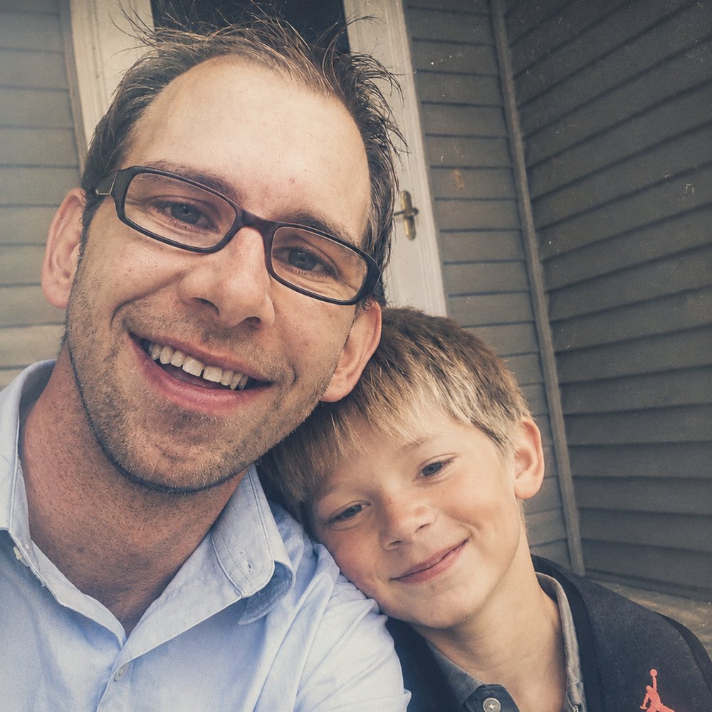 A man in glasses and a young boy leaning together and smiling outside a grey clapboard house