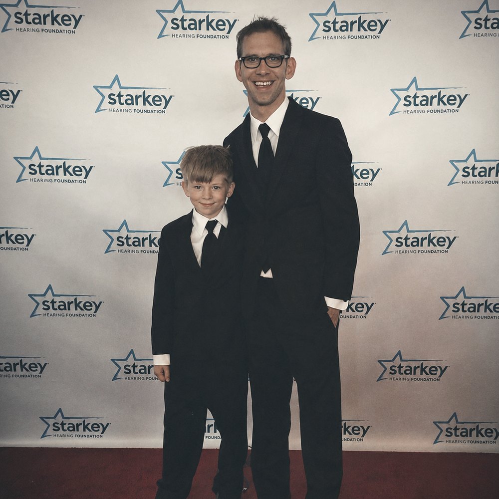 A man in glasses and a young boy, both in black suits, in front of a Starkey Hearing Foundation backdrop