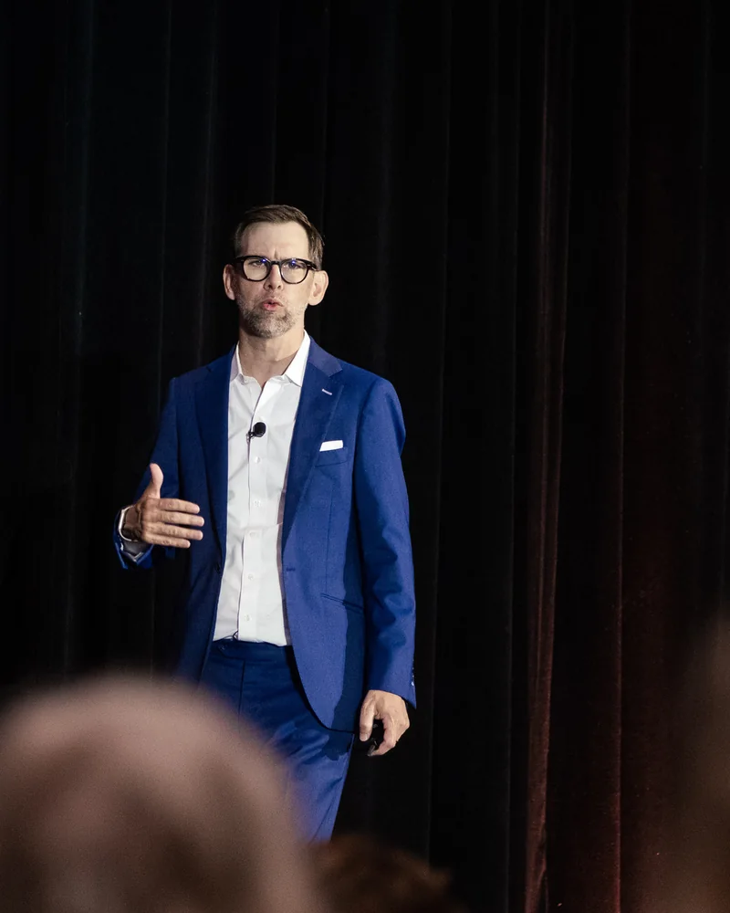 Michael Kutcher on stage in a navy suit, gesturing as he speaks