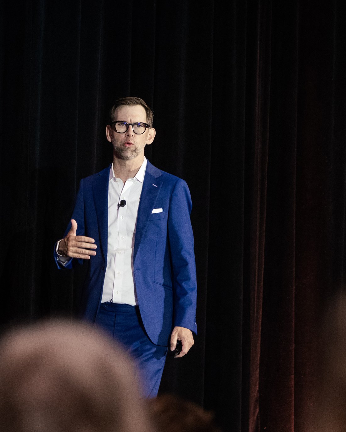 Michael Kutcher on stage in a navy suit, gesturing as he speaks